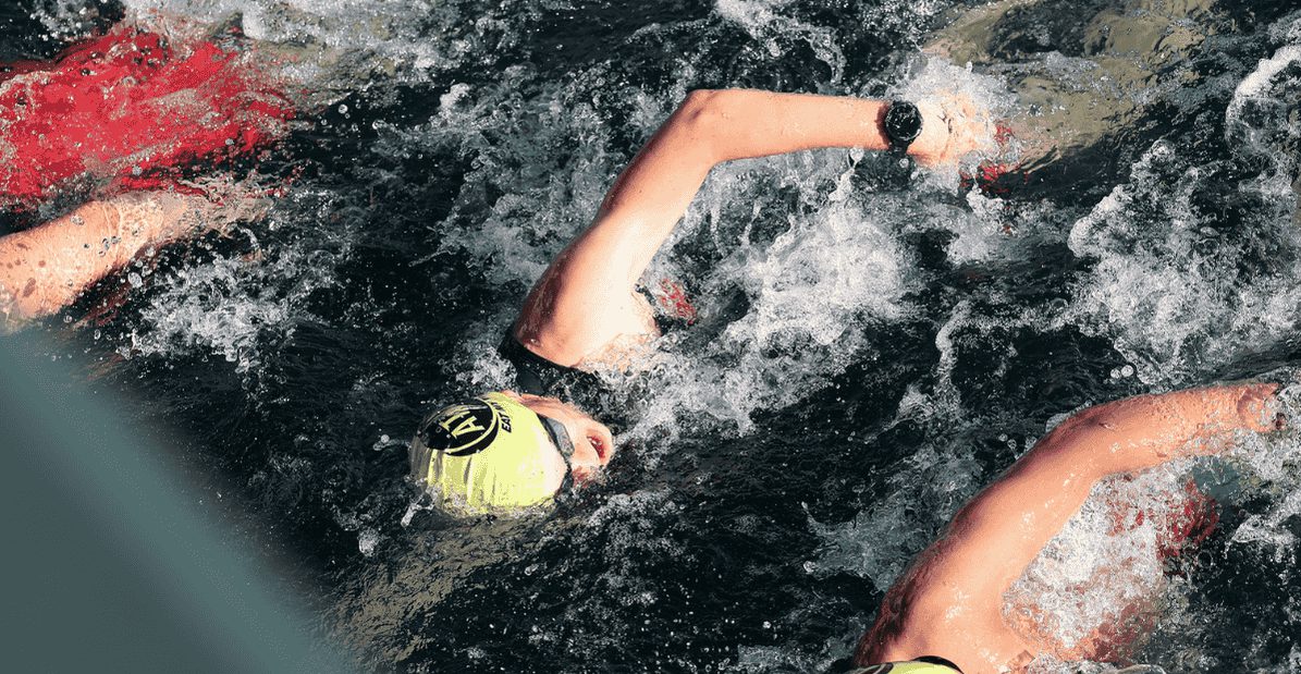 Two swimmers wearing yellow caps swim freestyle in choppy water, surrounded by splashes and ripples. One swimmer’s arm, wearing a watch, is extended forward above the water.