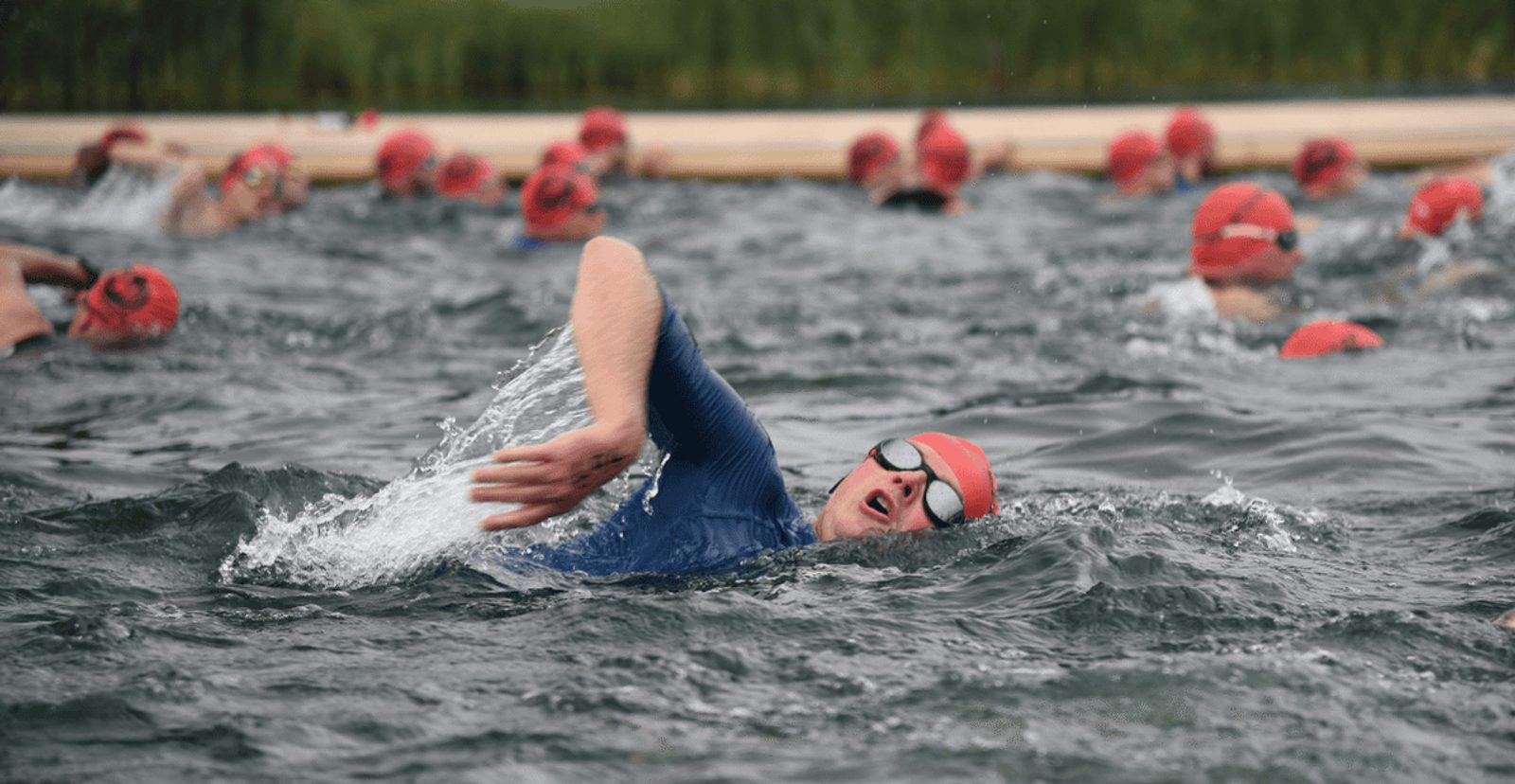 A group of swimmers wearing red swim caps and goggles participate in an open water race. One swimmer in the foreground performs a freestyle stroke, surrounded by choppy water, while others swim in the background.