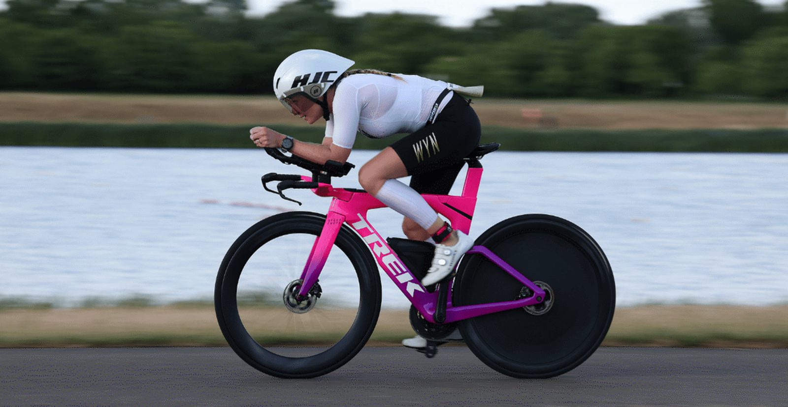 A cyclist wearing a white helmet and black-and-white cycling gear rides a bright pink Trek time trial bike on a paved road next to a river, with green trees in the background.