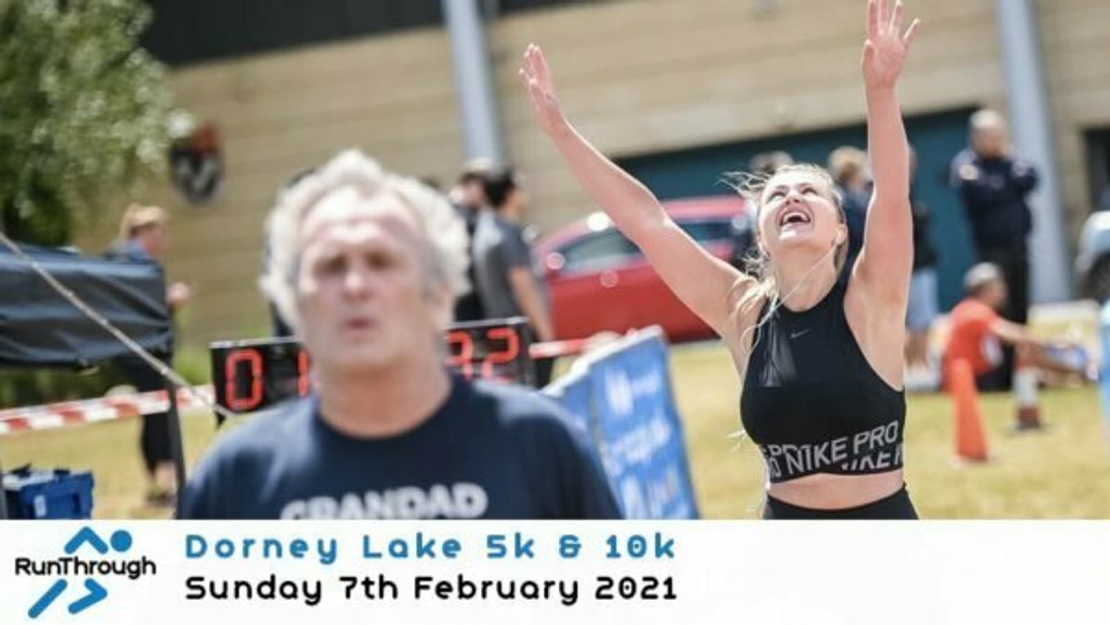 A woman is joyfully raising her arms at the finish line during a race at Dorney Lake, while a man jogs in front. The event is "Dorney Lake 5k & 10k" held on Sunday, 7th February 2021. A timer and signs are visible in the background.