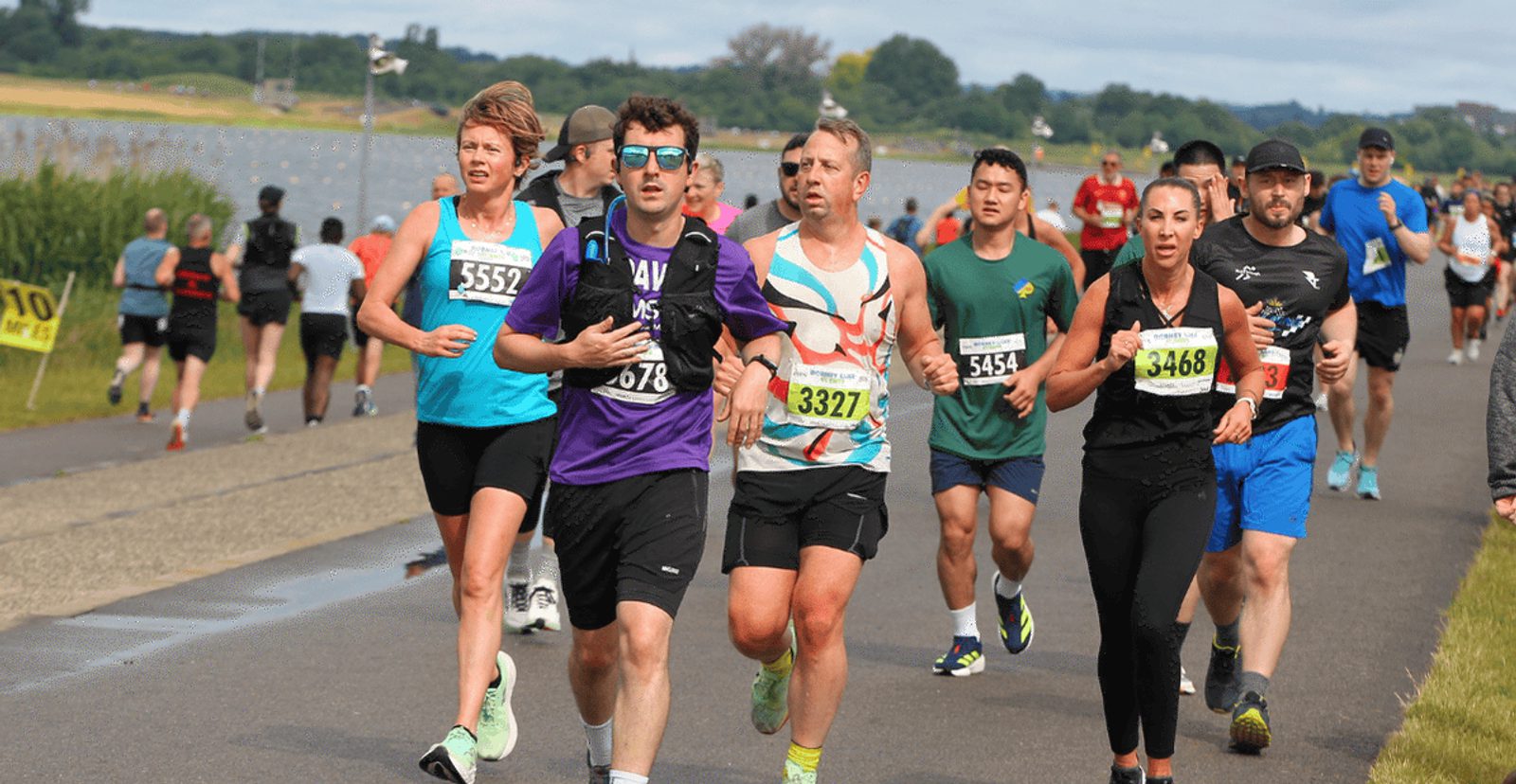 A group of runners with race bibs jog along a paved path near water and greenery on a sunny day. Some look focused while others chat. Additional runners are visible in the background.