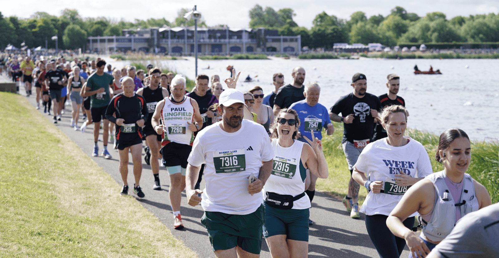 A large group of people run along a riverside path in a race, wearing numbered bibs. The weather is sunny, and trees, grass, and a building are visible in the background.