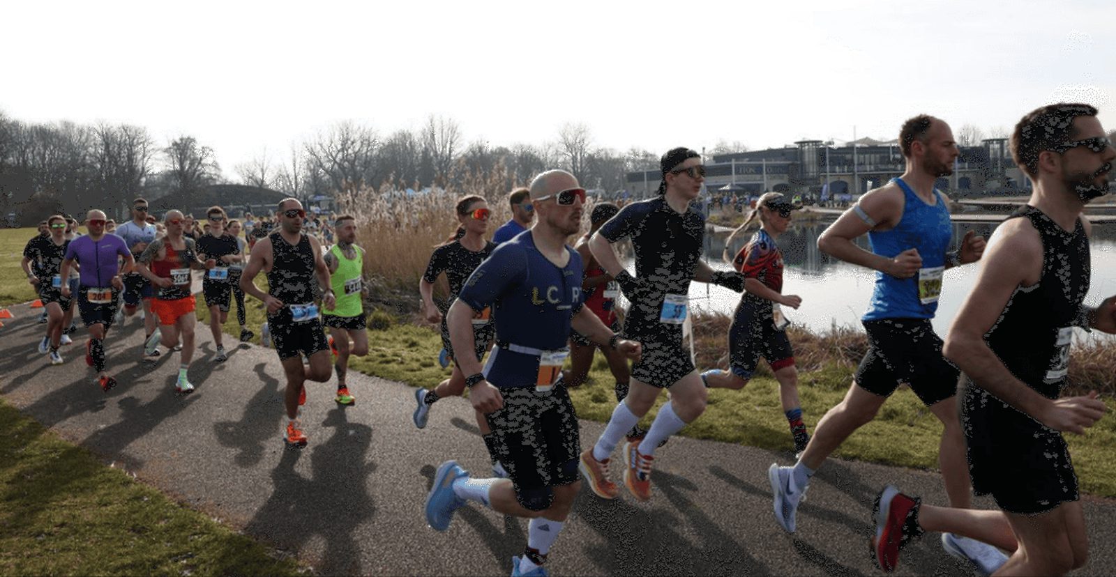A group of runners participate in a race along a lakeside path on a sunny day, with trees and buildings visible in the background.