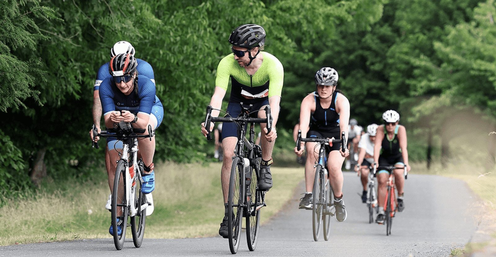 A group of cyclists ride on a paved road surrounded by greenery. The front riders wear helmets and athletic gear, with others following closely behind on a sunny day.