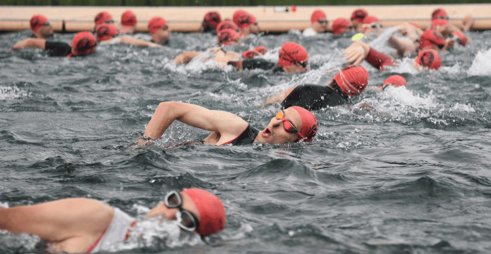 A group of swimmers wearing red swim caps and goggles compete in an open water swimming race, creating splashes as they move through the water.