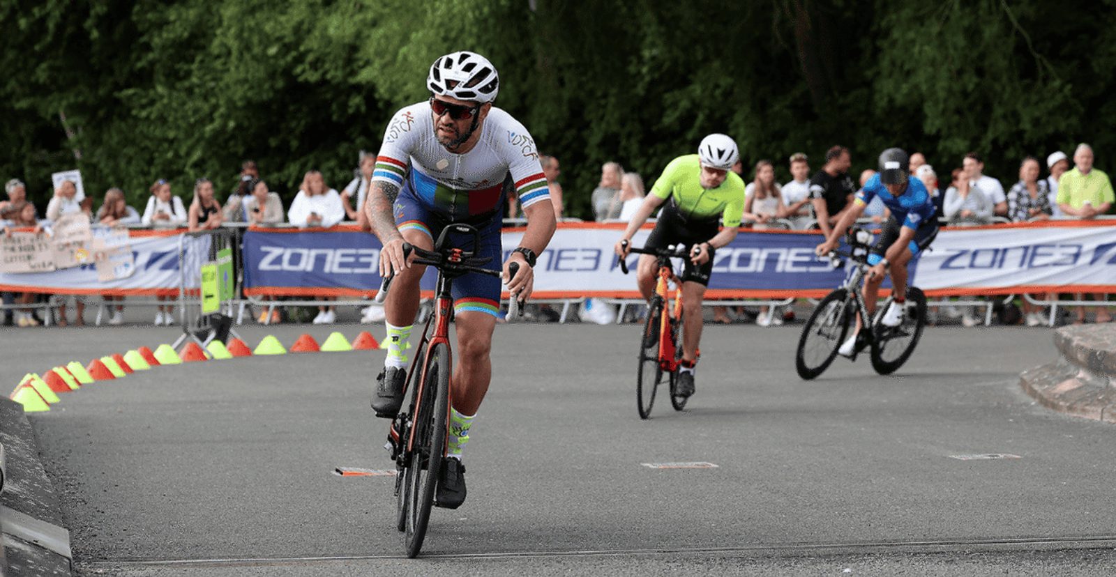 Three cyclists race on a paved road during a competition, watched by spectators behind barriers. The leading cyclist wears a white jersey with colorful stripes and a helmet, while the others follow behind. Trees are in the background.