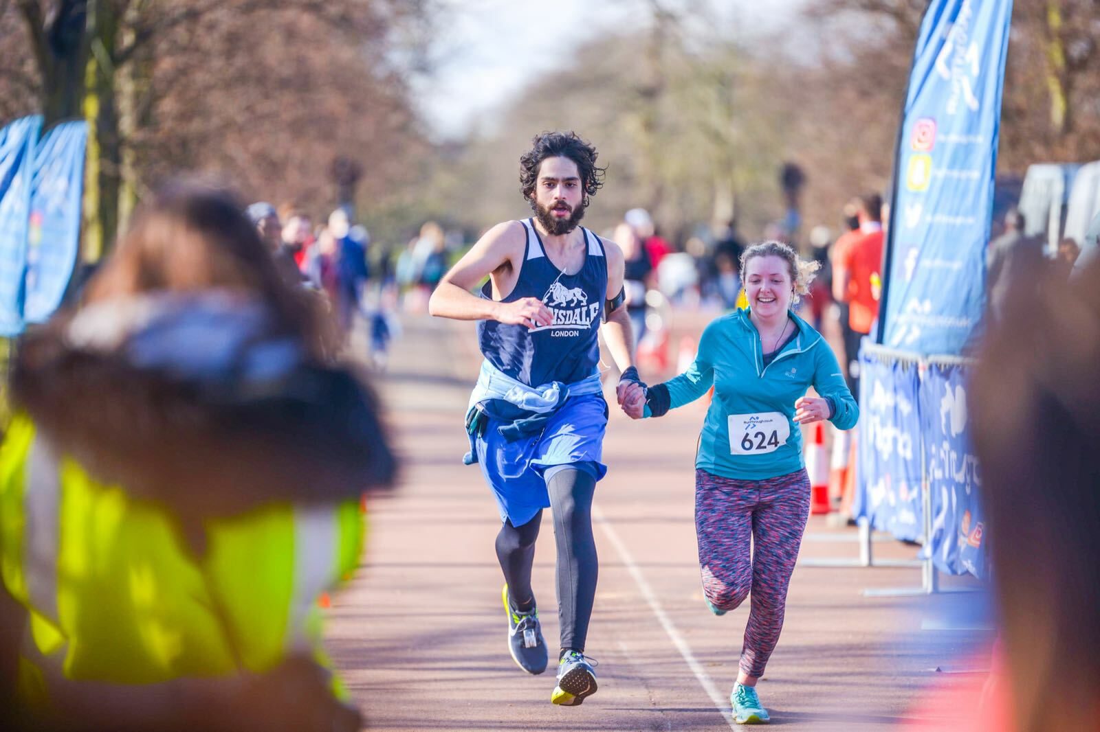 Two runners, a man and a woman, race toward the finish line in a marathon event. The man wears a dark blue tank top and shorts, and the woman wears a light blue top and colorful leggings. They are both smiling and holding hands. Other runners and spectators are in the background.