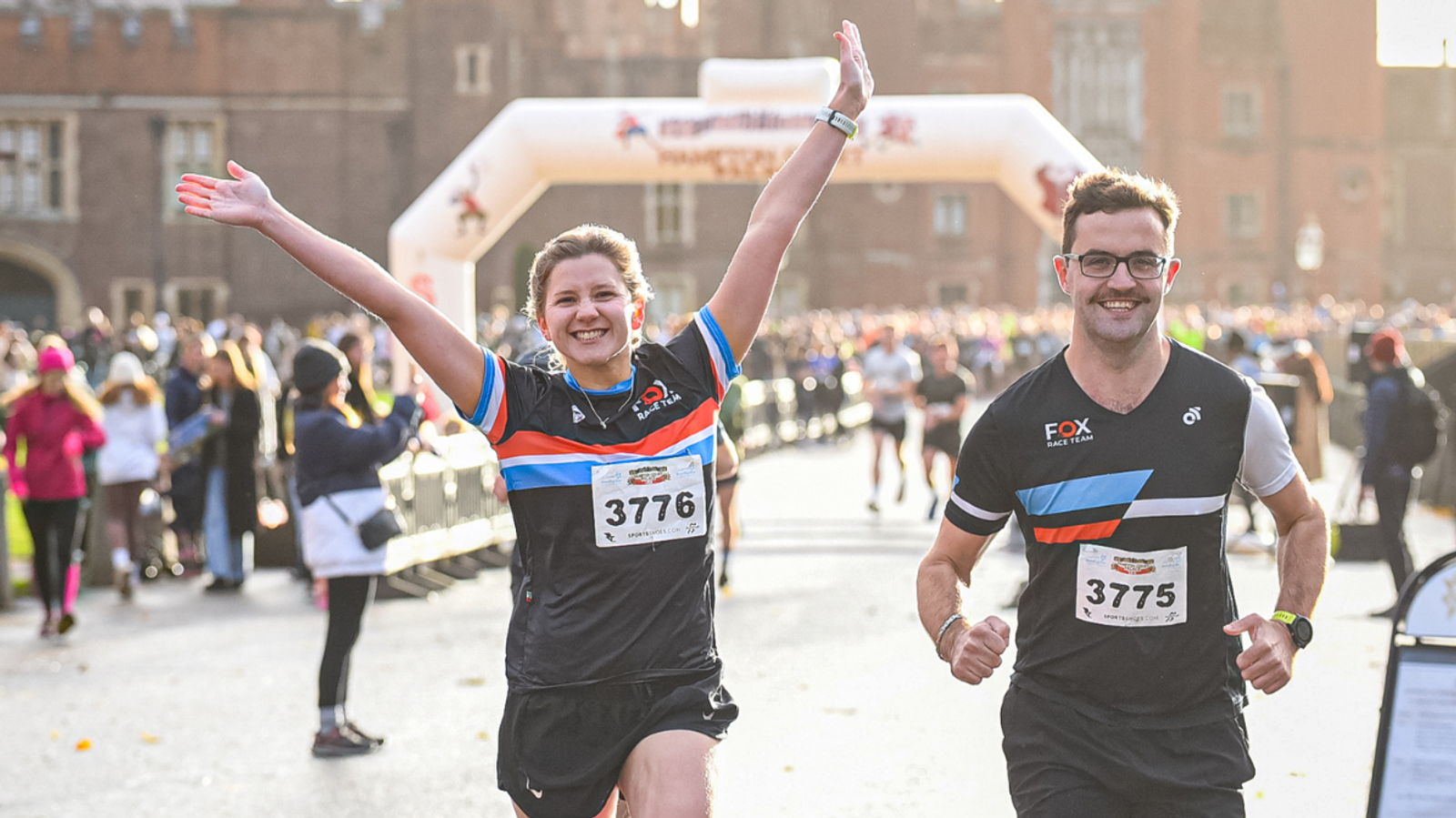 Two runners with race numbers 3776 and 3775 smile and raise their arms celebrating as they cross the finish line in a crowded outdoor race. The bright day shows many other participants behind them.