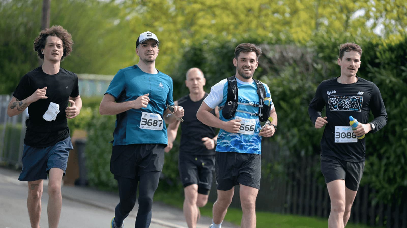 Five runners on a road, with race bib numbers visible, running outdoors. They appear focused, dressed in athletic gear. Greenery and a fence are in the background.