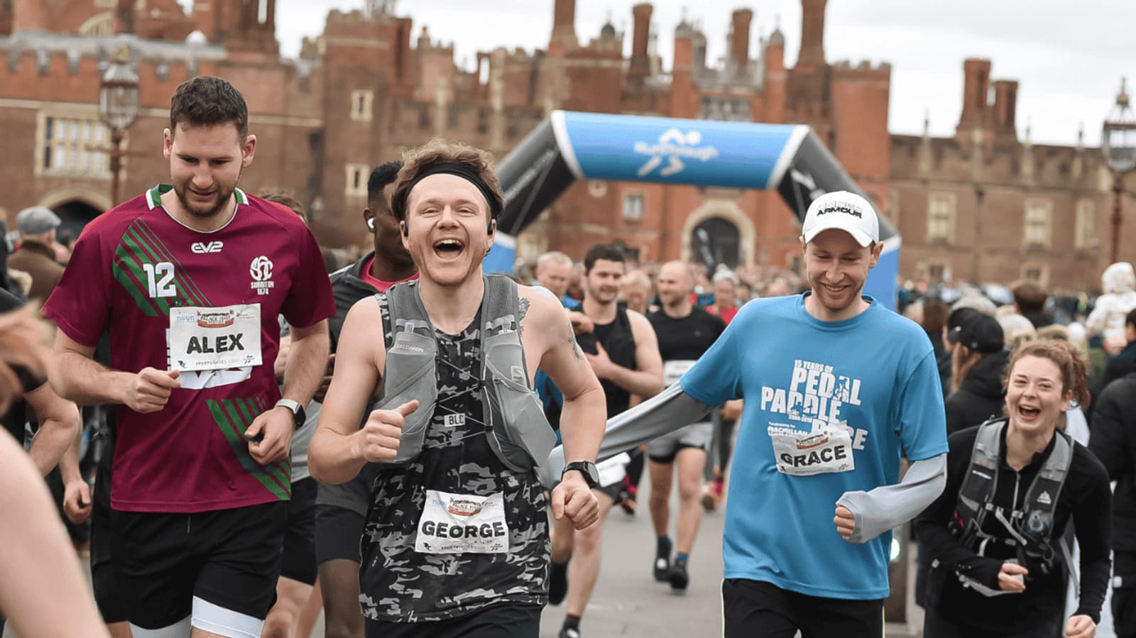 A group of runners, smiling and wearing race bibs, participate in an outdoor race with a large historic building and a blue inflatable arch in the background. The atmosphere is lively and energetic.