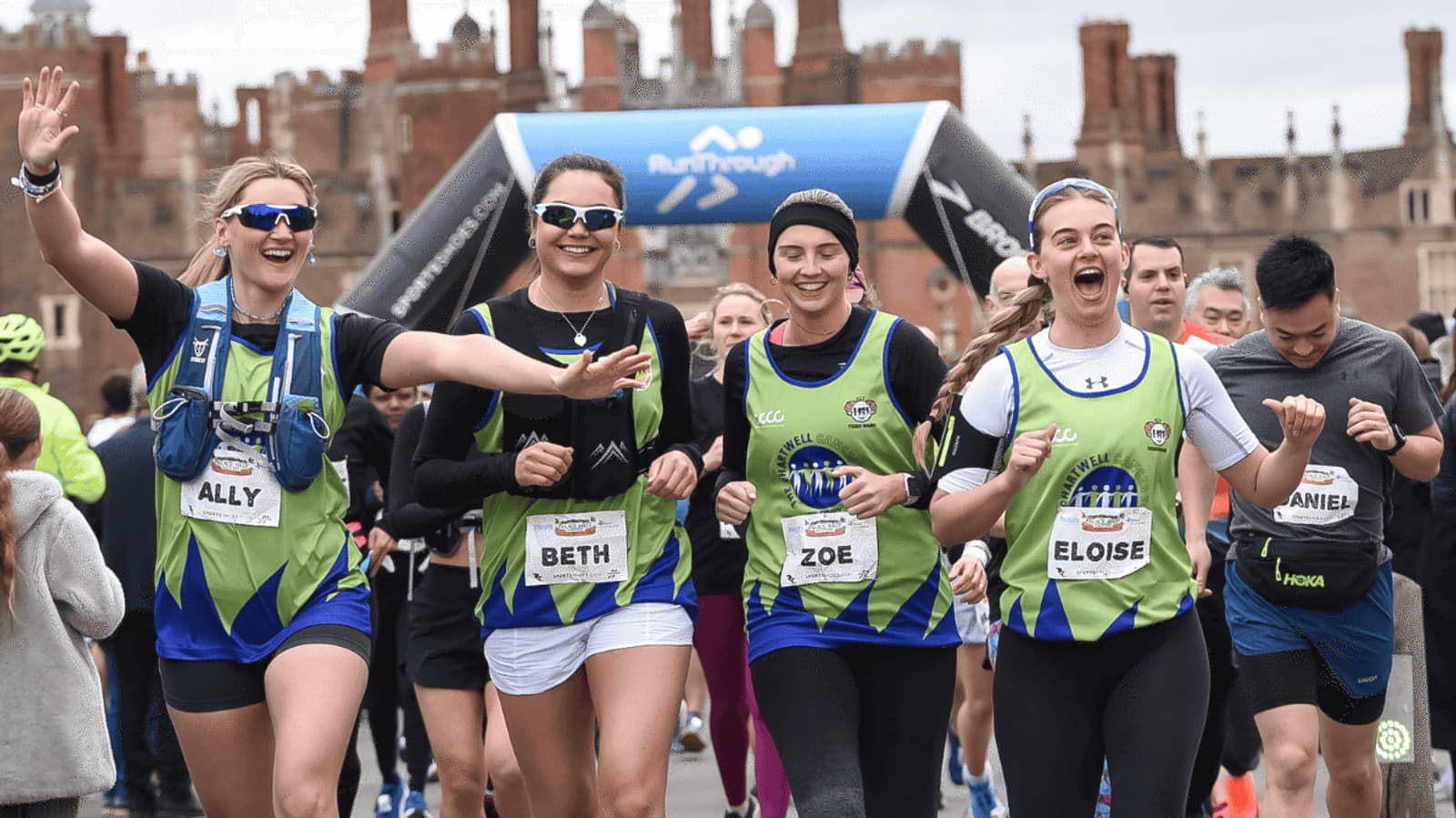 Four women in matching green and blue running outfits smile and cheer as they run in a race. Other runners are around them, and a historic building is visible in the background.