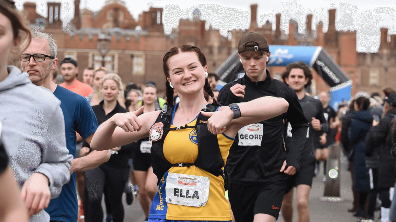 A smiling runner in a yellow and black vest labeled "Ella" gives double thumbs up during a race, surrounded by other runners. Historic brick buildings are visible in the background.