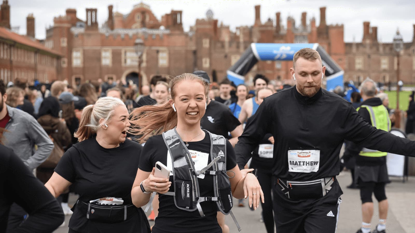 A group of runners, wearing race bibs and hydration packs, smile and run in an outdoor event near a historic brick building. The atmosphere is lively, with many participants and supporters in the background.