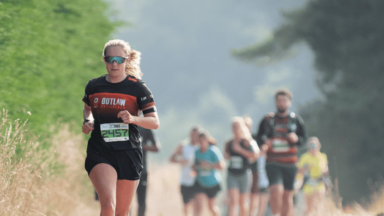 A woman wearing sunglasses and a black "Outlaw" shirt runs a race on a path with grass on the side. Other runners follow behind her, slightly out of focus. She has a race bib numbered 2457.