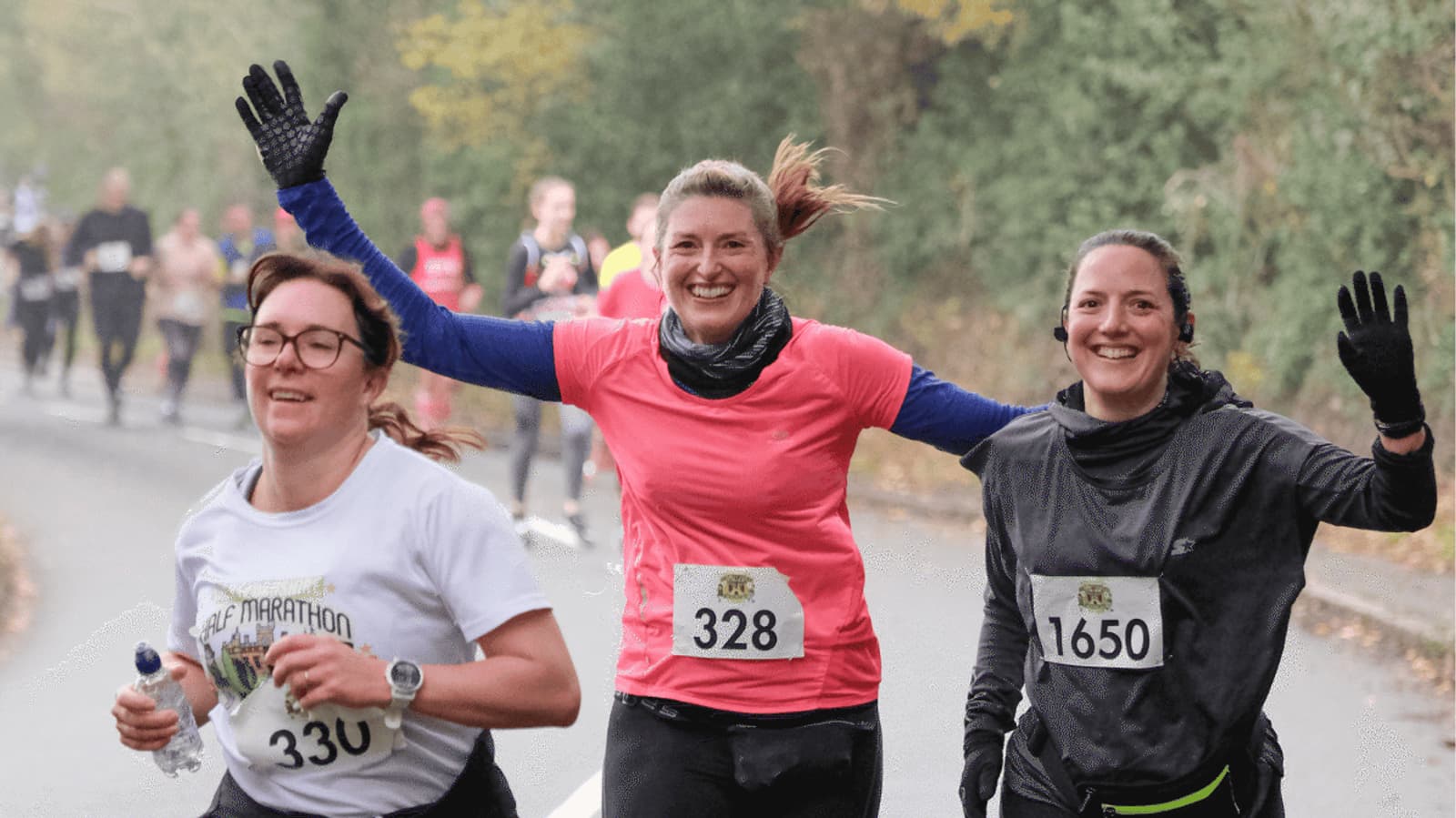 Three women are running in a race on a road. The woman in the center, wearing a pink shirt and bib number 328, smiles with arms raised. Other runners can be seen in the background.