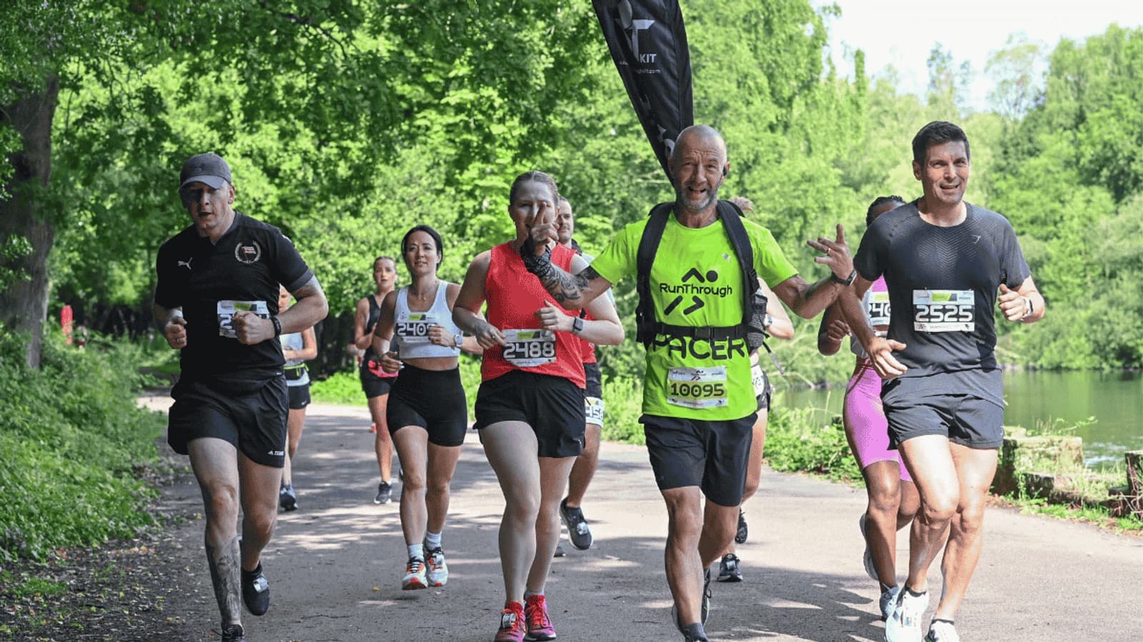 A group of runners, wearing race bibs, run along a tree-lined path next to a lake on a sunny day. The participants look energetic and focused.