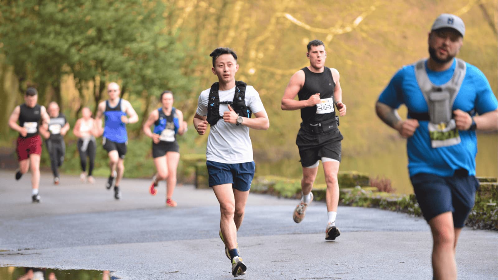 A group of runners participate in a race along a paved path beside trees and water on a sunny day, with some runners in the foreground and others in the background.