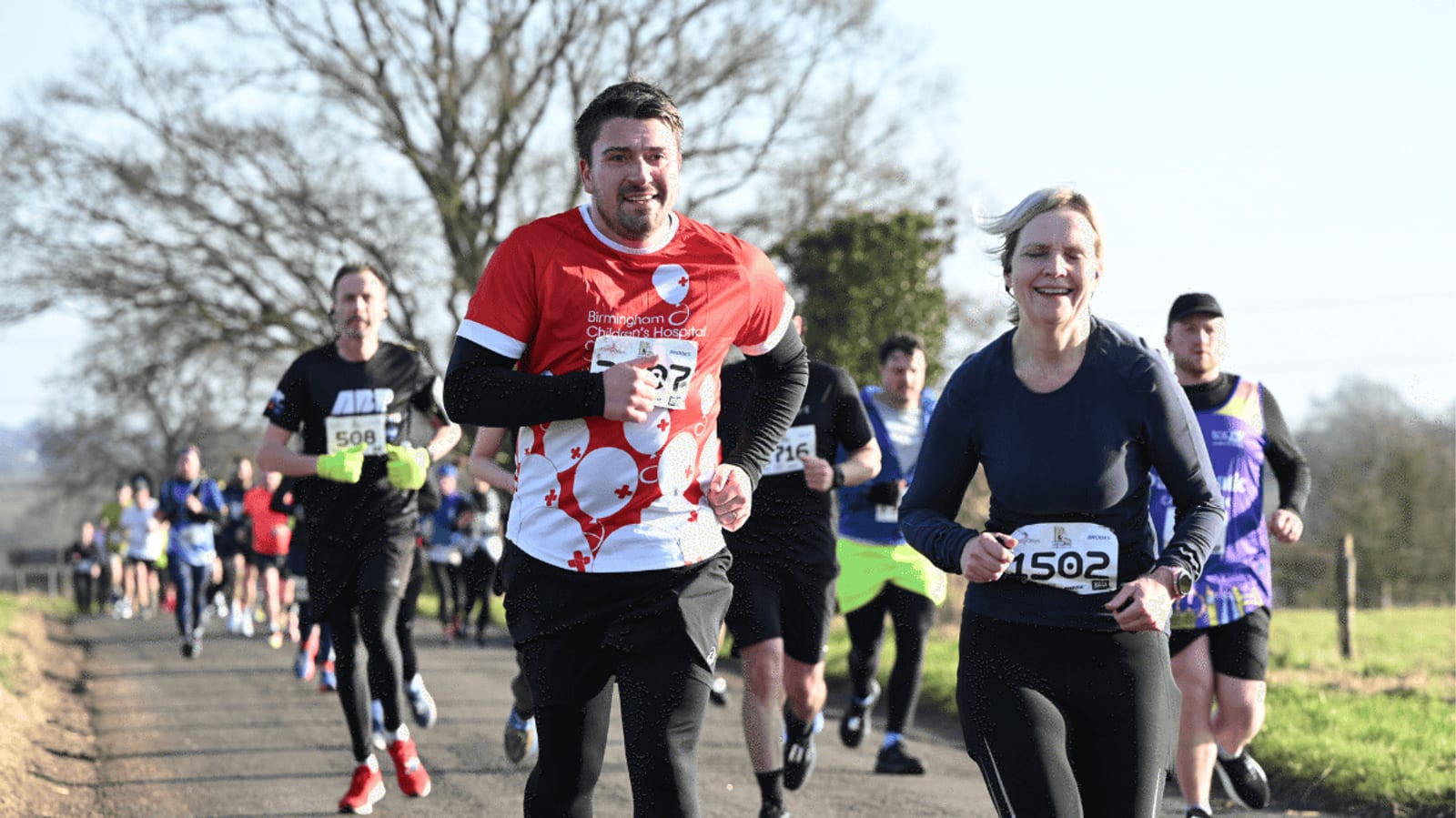 A group of runners, wearing numbered bibs, participate in an outdoor race on a sunny day. Two runners, a man and a woman in front, smile as they run along a rural road with others following behind.