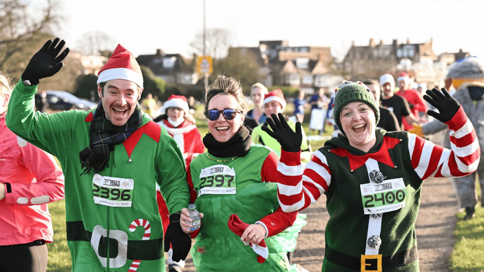 Three people in festive holiday costumes smile and wave while running in a race. They wear Santa hats, elf outfits, and race bibs, surrounded by other cheerful participants outdoors on a sunny day.