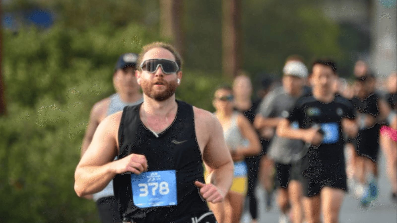 A man wearing sunglasses and a black tank top runs in a race with a blue bib numbered 378, followed by a group of runners on a sunny outdoor course.