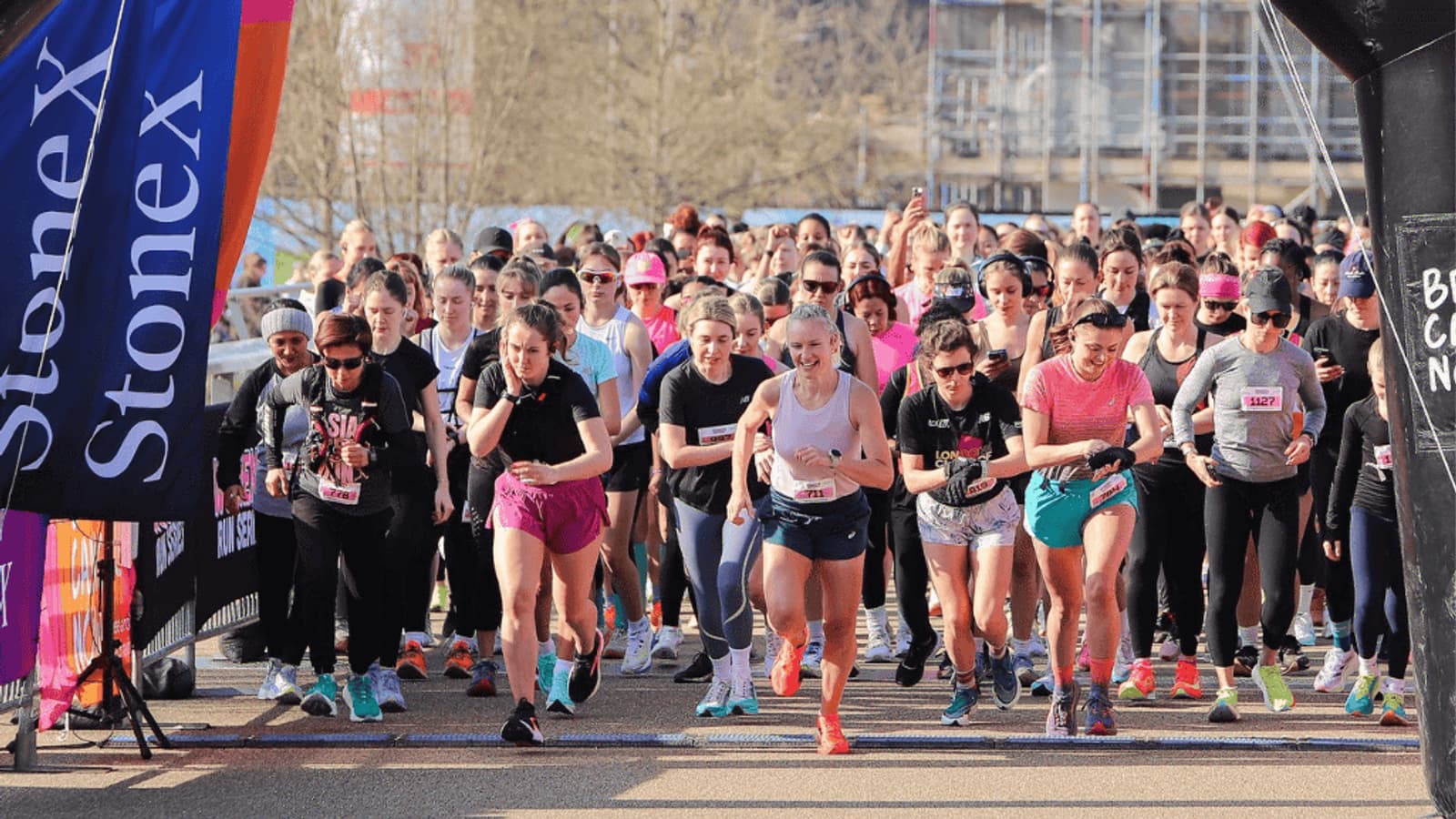 A large group of women runners start a race outdoors on a sunny day, with a "Stonex" banner visible on the left and most participants wearing athletic clothing and running shoes.