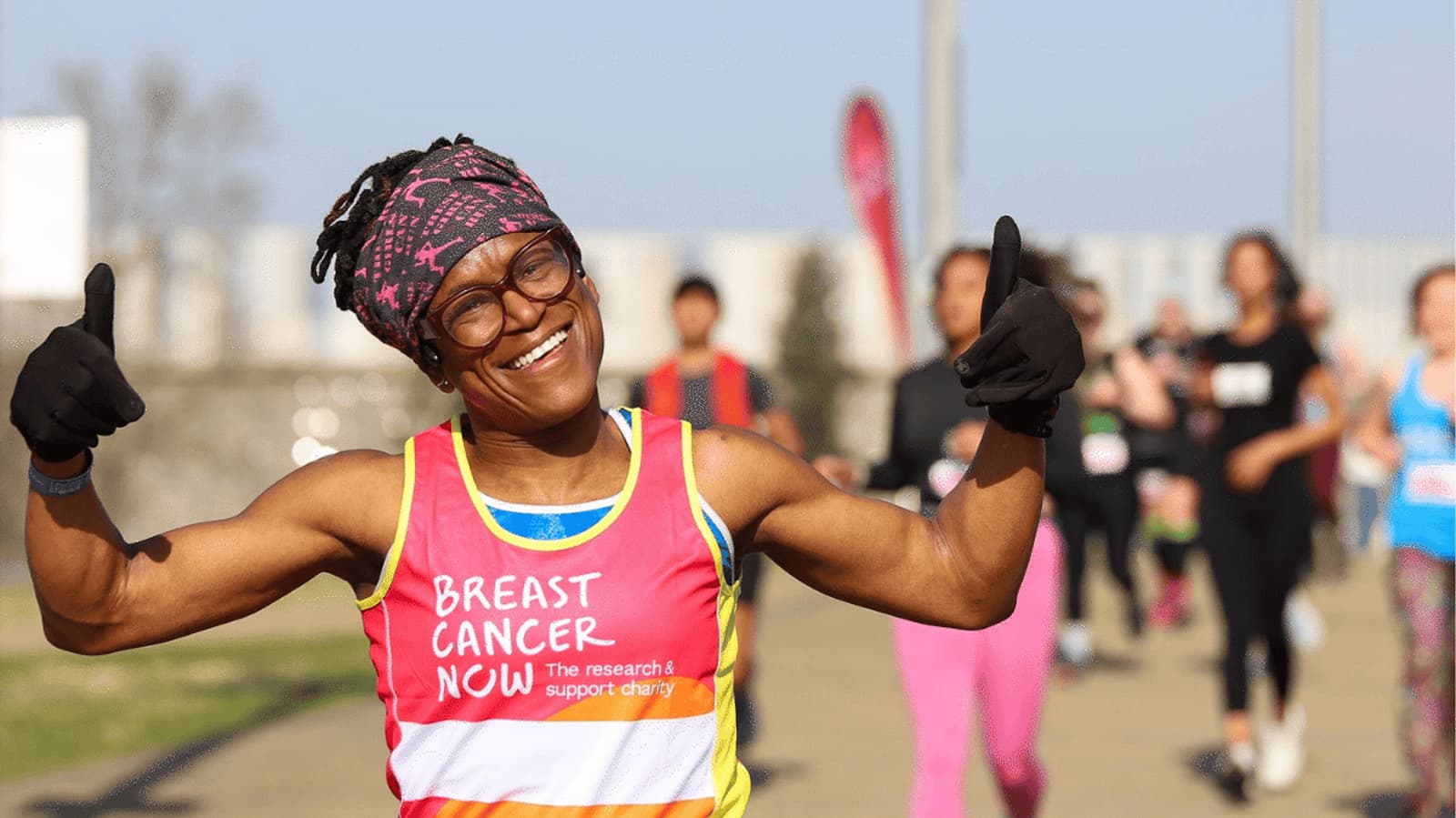 A smiling runner wearing a "Breast Cancer Now" tank top and a headband gives two thumbs up during a race, with other participants visible in the background.