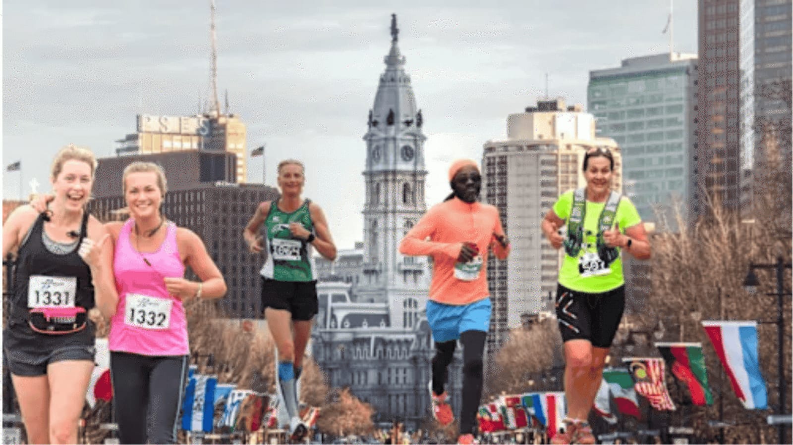 Five marathon runners smiling as they run through a city street lined with flags, with Philadelphia’s City Hall and skyscrapers visible in the background.