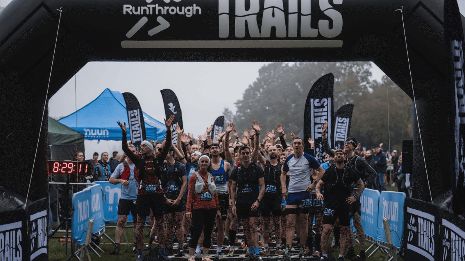 A group of runners gather excitedly at the starting line under a black inflatable arch labeled "RunThrough TRAILS," with hands raised and race banners and timing equipment visible, ready for a trail race.