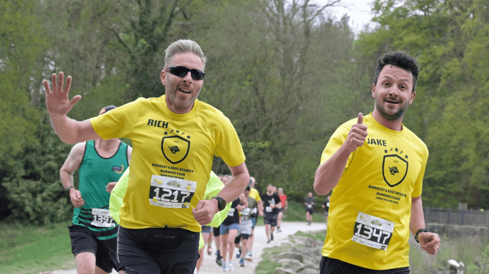 Two men in yellow shirts labeled "RICH" and "JAKE" run a race outdoors, smiling and waving at the camera. Other runners in various colored shirts follow behind them on a gravel trail lined with trees.