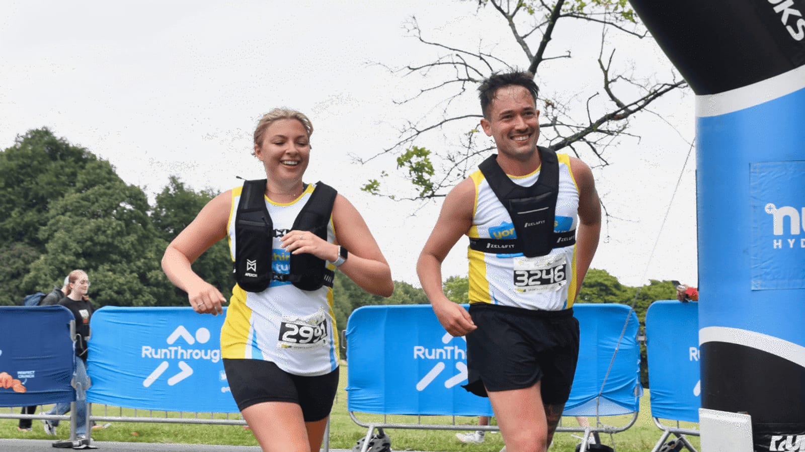 Two smiling runners in race bibs, wearing hydration vests, cross a finish line outdoors with blue barriers and trees in the background.