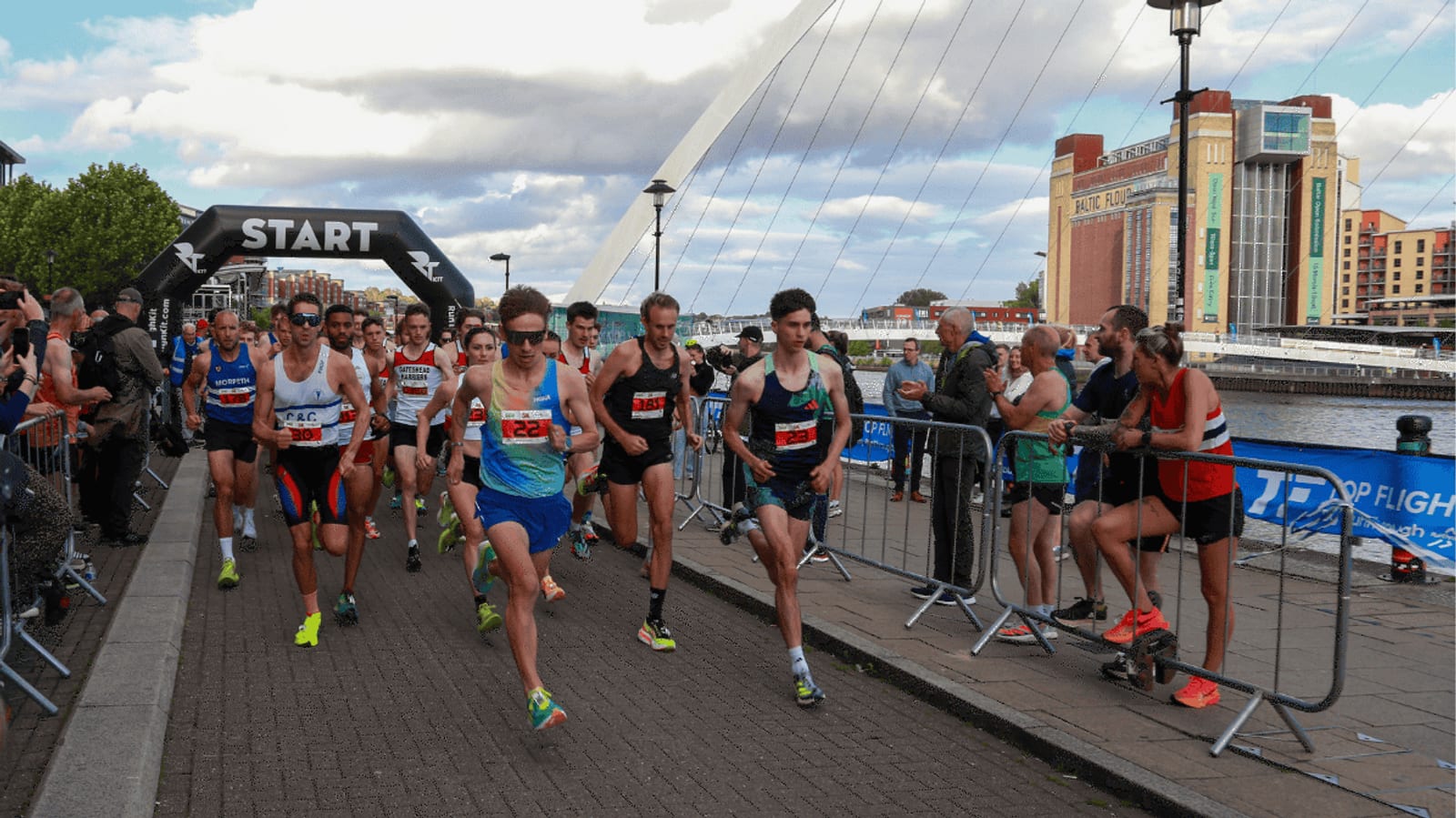 A group of runners wearing race bibs start a marathon near a waterfront, as spectators watch and photographers take pictures. A large "START" banner and a modern bridge are visible in the background.