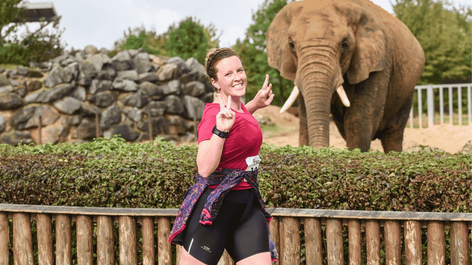 A woman in athletic clothing runs past a wooden fence, smiling and making peace signs with both hands; an elephant stands behind her on the other side of the fence, with greenery and rocks in the background.