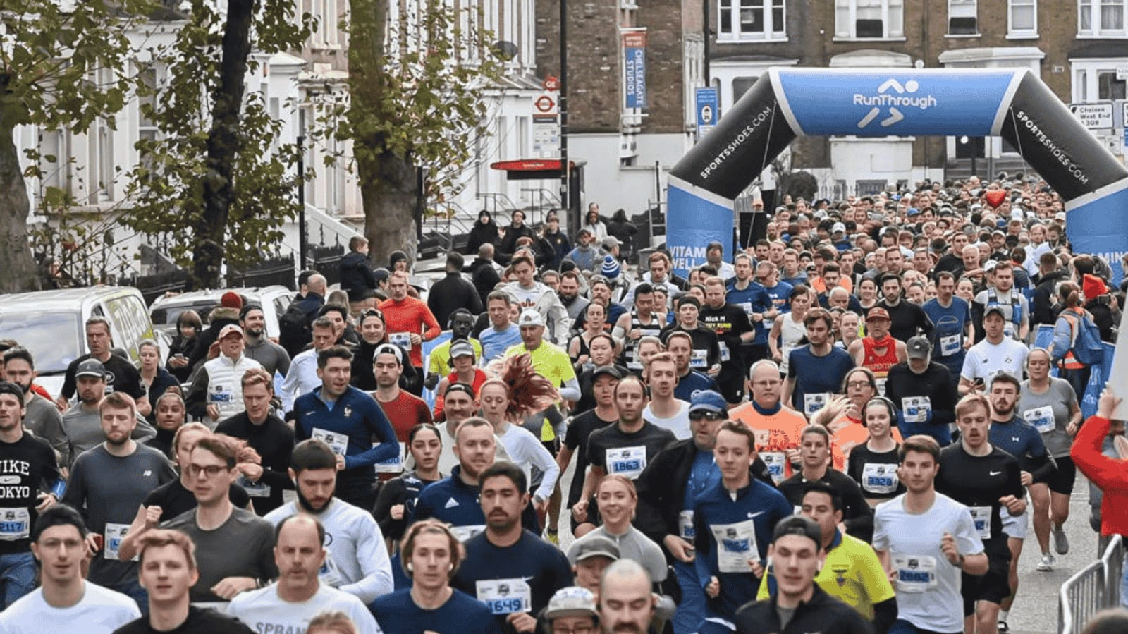 A large crowd of runners participate in a road race, passing under a blue inflatable archway labeled “RunThrough.” Trees and buildings line the street, and many participants wear numbered bibs and athletic clothing.