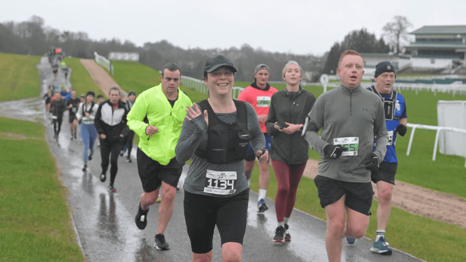 A group of runners participate in a race on a wet, paved path. The leading woman, smiling, wears a cap and race bib 1134. Other runners in various athletic outfits follow close behind under a gray, overcast sky.