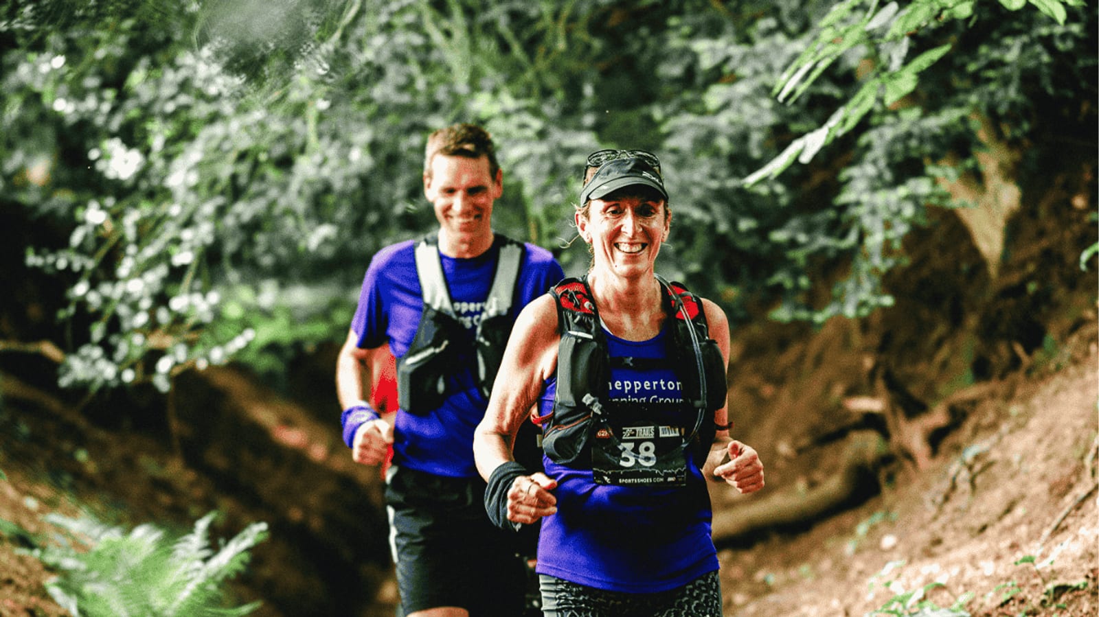 Two smiling runners in purple shirts and race bibs run on a forest trail, surrounded by greenery. The woman is in front, and both appear to be enjoying the outdoor activity.