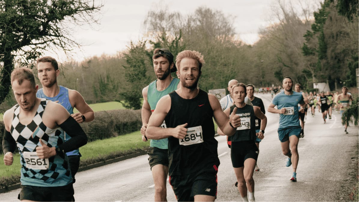 A group of runners, wearing numbered bibs, participate in a road race on a tree-lined street. The lead runner smiles and gives a thumbs up, while others run closely behind him. The weather appears overcast.