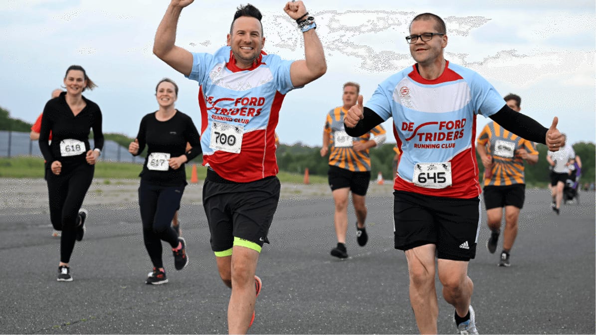 Two smiling runners wearing "De Ford Striders Running Club" shirts and numbered bibs raise their arms in victory as they lead a group of other runners on an outdoor road during a race.
