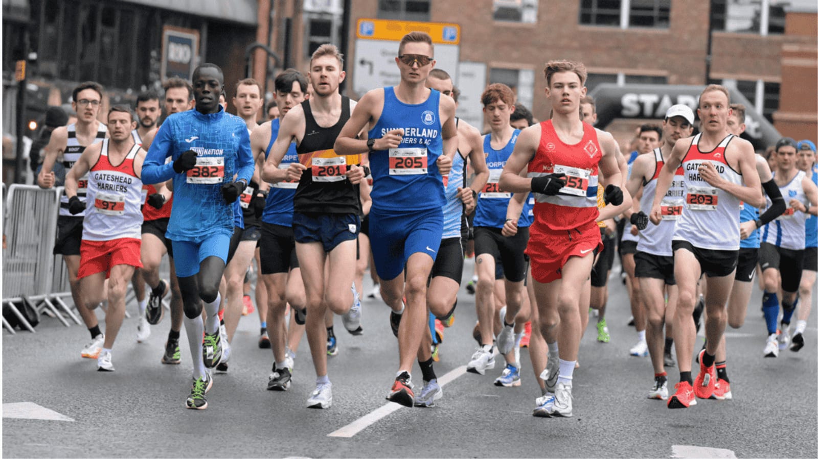 A group of runners wearing race bibs compete in a road race on a city street, with some in blue and others in red or white uniforms. Buildings and spectators are visible in the background.