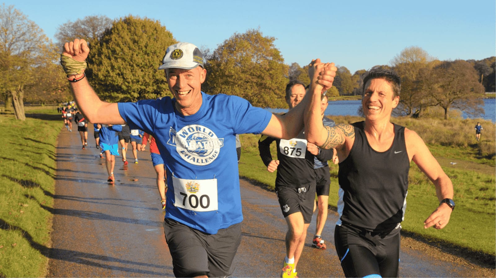 Two runners, smiling and holding hands in celebration, cross a finish line during a race on a sunny day. Both wear numbered bibs and athletic gear; other runners and trees are visible in the background.
