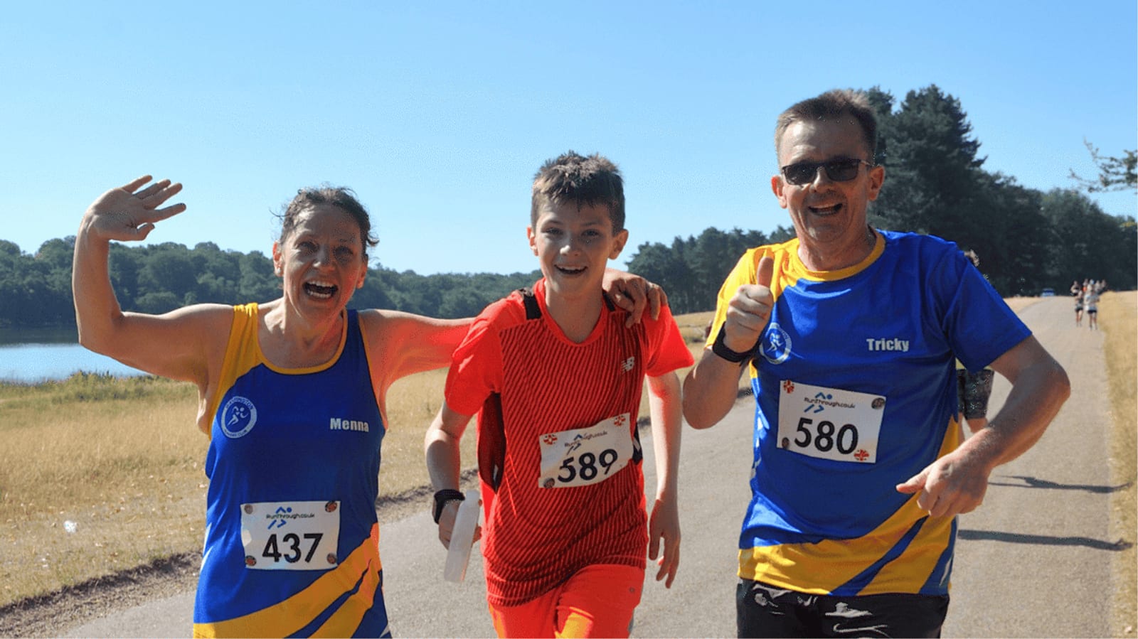 Three smiling runners, two adults in matching blue and yellow shirts and a boy in red, run together on a sunny path by a lake, with trees and clear blue sky in the background.