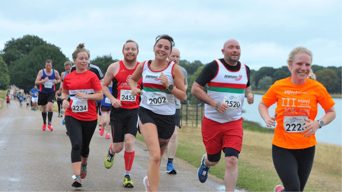A group of runners, wearing race bibs and athletic gear, smile as they participate in a road race on a cloudy day. Trees and grass line the path, and a lake is visible in the background.