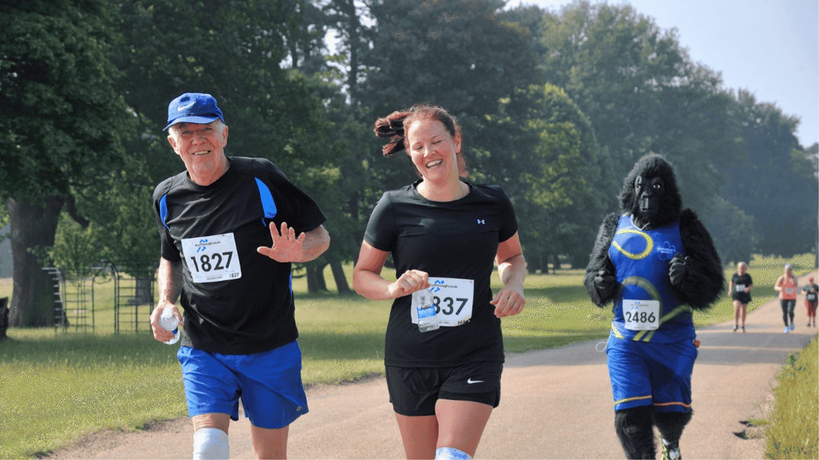 Three people are running a race outdoors: two smiling runners in athletic gear and one person dressed in a gorilla costume, all wearing race numbers. Other participants are visible in the background.
