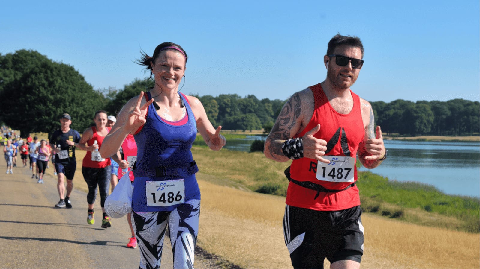 Two runners, a woman and a man, smile and give thumbs up as they participate in a race by a lake on a sunny day. Other runners follow behind on the path, surrounded by grass and trees.