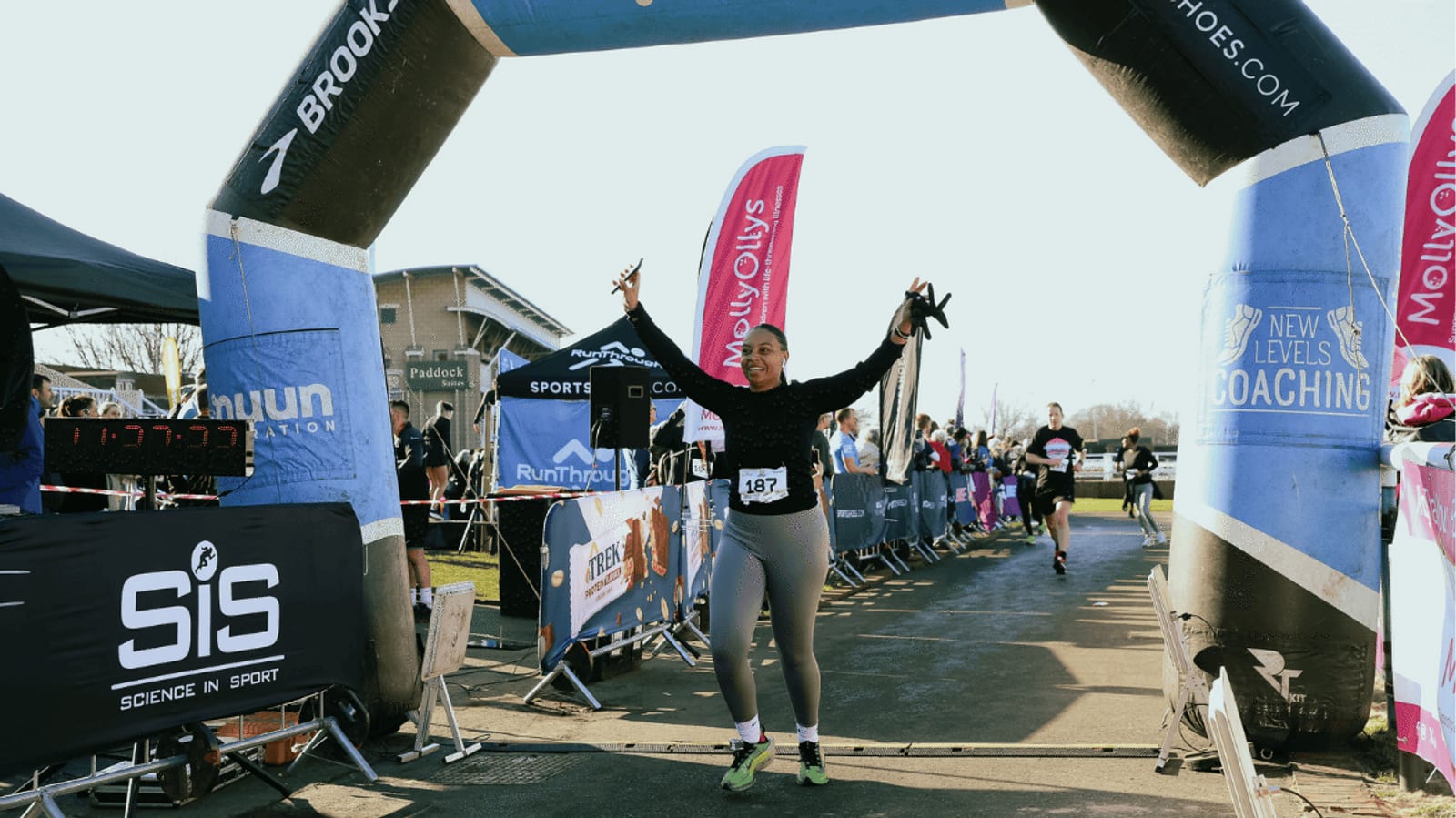 A woman smiles and raises her arms in celebration as she crosses the finish line at a race event, with other runners and spectators visible in the background under a large inflatable arch.