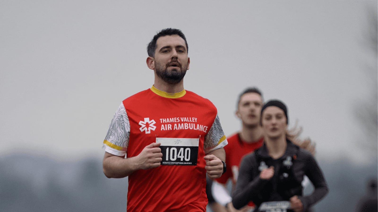 A man wearing a red "Thames Valley Air Ambulance" shirt and race bib 1040 runs outdoors, with three other runners visible in the blurred background. The sky appears overcast.