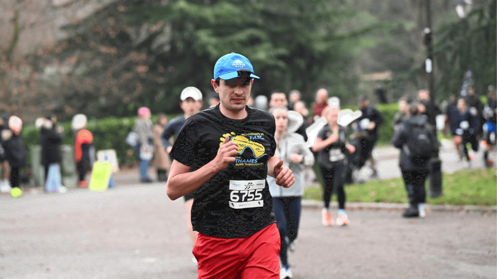 A man in a blue hat and black shirt with a race bib runs in an outdoor race. Several other runners and blurred spectators are in the background, with trees and greenery lining the park path.