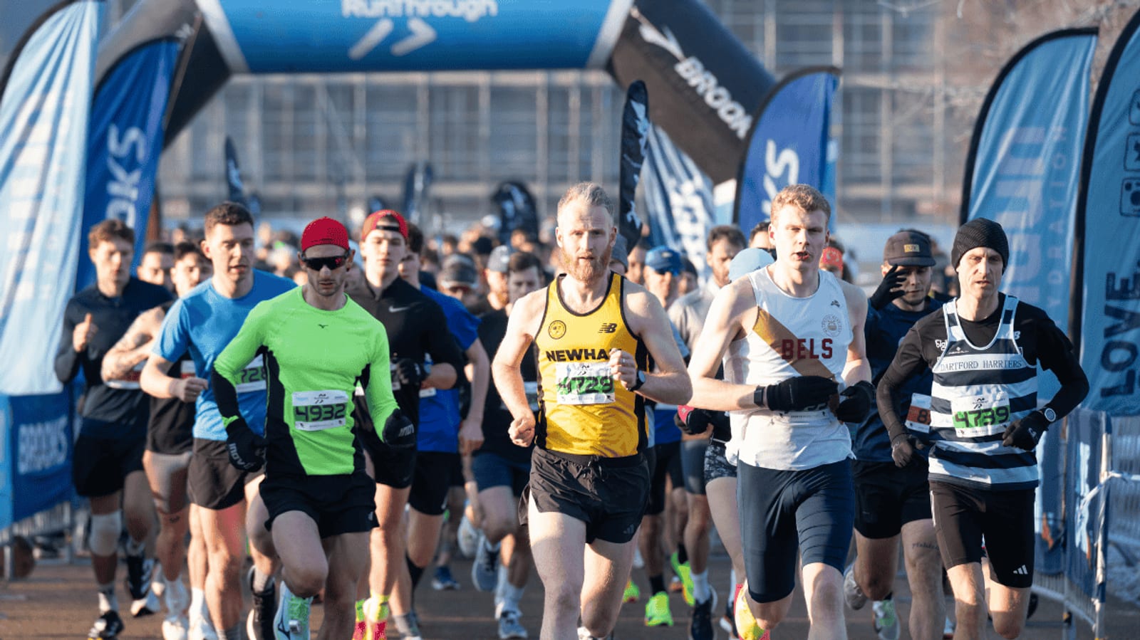 A group of runners in athletic gear compete in a road race, approaching the starting line under a blue RunThrough archway on a sunny day.
