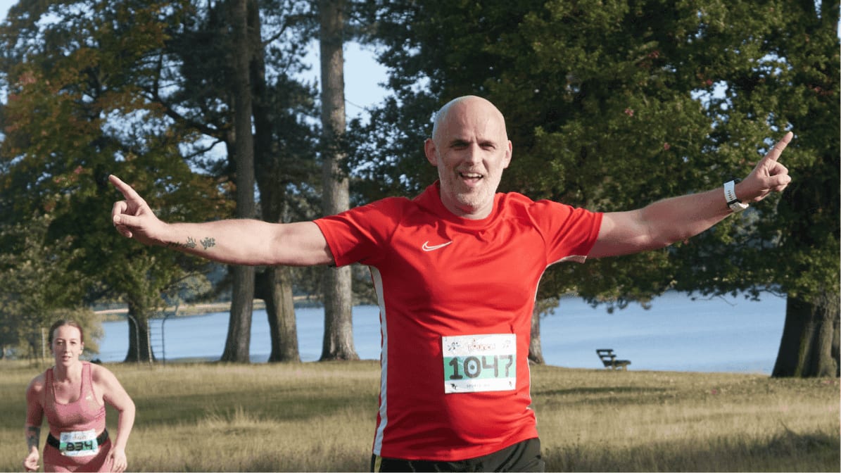 A man in a red shirt with race number 1047 runs outdoors, smiling with arms raised in celebration. A woman follows behind him; trees and a lake are visible in the background.