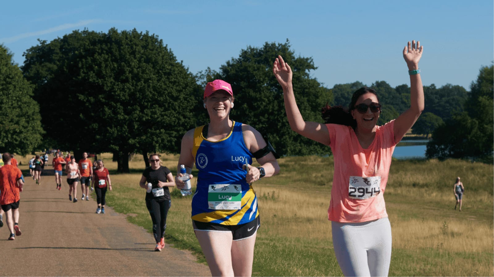 Two women are running in a race on a sunny day. One wears a blue and yellow vest and pink hat, smiling, while the other in a pink shirt raises her arms in celebration. More runners and green trees are visible in the background.