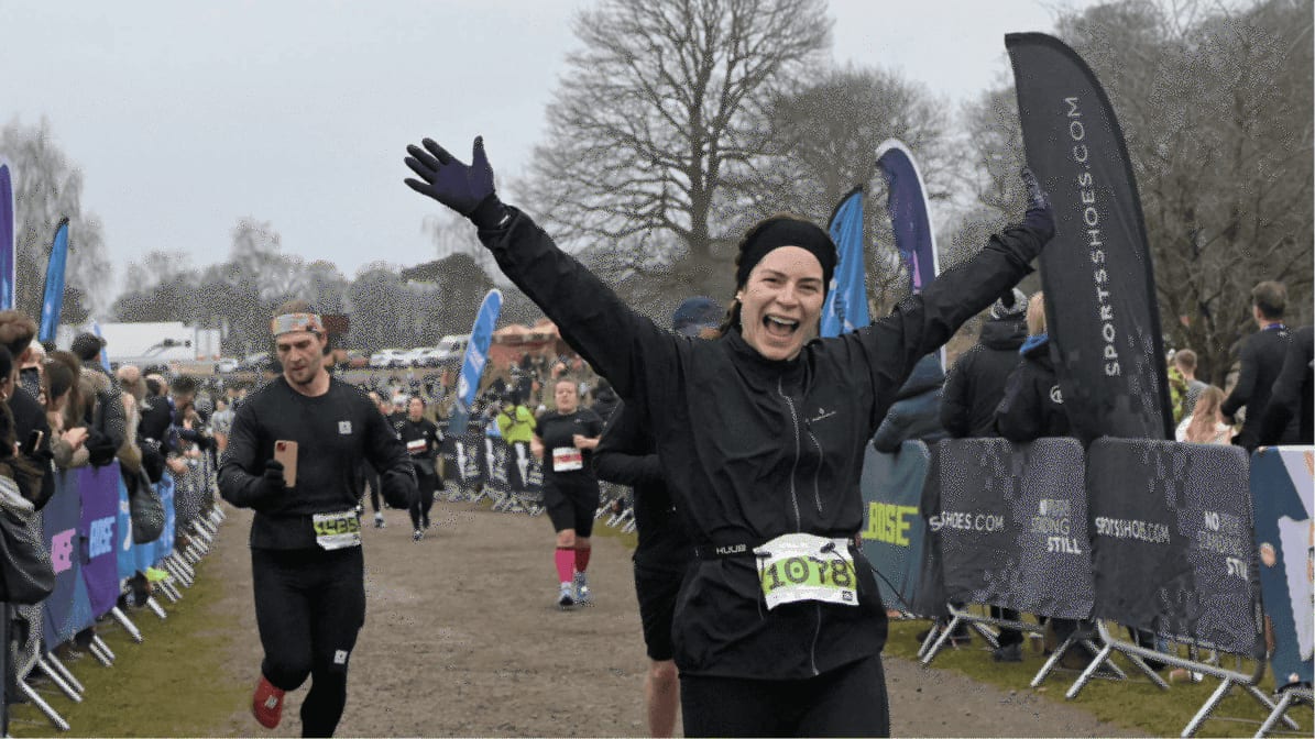 A woman wearing a black outfit and green race bib 1078 smiles with arms raised as she crosses a race finish line, with other runners and cheering spectators behind barriers on a cloudy day.
